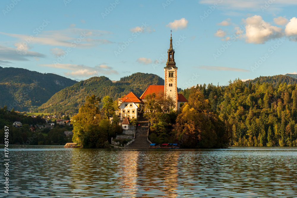 Fototapeta premium Peaceful sunset view of Lake Bled with the island church and castle reflecting in calm waters, surrounded by soft autumn light and mountain silhouettes