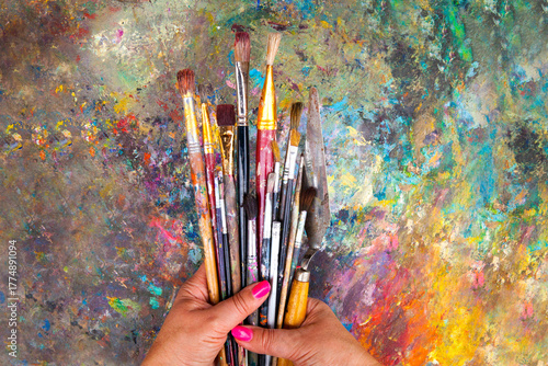 Artist holding a bunch of used paintbrushes over a colorful abstract background. Creative workspace, art inspiration, painting tools, and artistic expression concept.