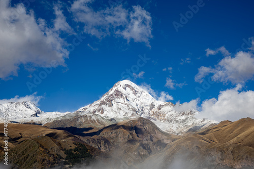 A breathtaking view of the Mount Kazbegi, featuring its snow-covered summit sharply contrasted against a deep blue sky and surrounded by dry, rocky mountain slopes and patches of low-hanging mist