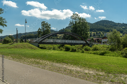 Modern Arch Footbridge in Muszyna, Poland, with the sunny Beskid Sadecki Mountains in the background.