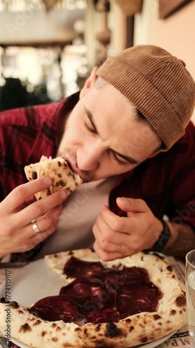 Young man eating pizza with prosciutto while sitting at cafe table in city center. Vertical 4k footage