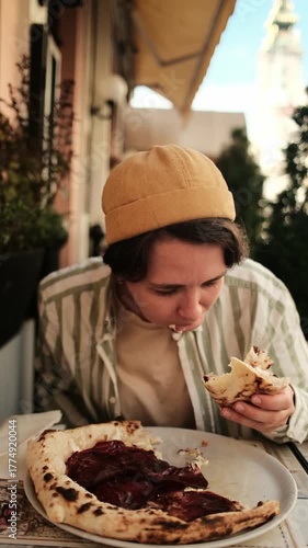 Woman with yellow cap eating slice of pizza with prosciutto while sitting at outdoor cafe on sunny day. Vertical 4k footage