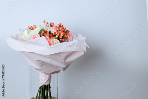 Elegant bouquet of white hydrangeas, pink carnations, and red berries wrapped in pastel pink paper, placed in a glass vase
