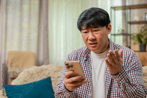 Papier peint Asian man sitting on home sofa reads bad fail news on smartphone, frowning, showing disappointed frustrated mood