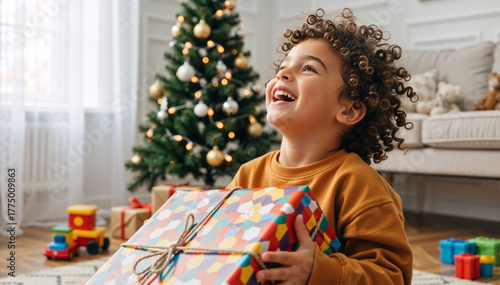 Happy little boy with curly hair laughing while holding a Christmas gift. Excited child receiving a present at home by the festive tree. Holiday season joy and childhood happiness