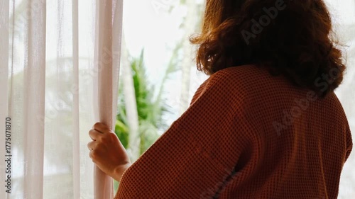 The woman was at the window in the bedroom. She opened the curtains on the window. In the morning and she looks at the view of mountains and trees at sunrise.	