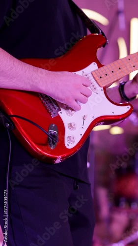 Hands strumming red electric guitar with steady rhythm under purple lights, musician wearing dark shirt, close framing on body and strings, stage ambience in background