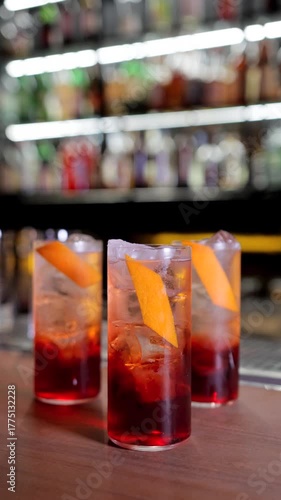 Three red negroni cocktails on bar counter, precise orange peel placement with tweezers, sparkling ice and shelf of bottles in background, elegant aperitif moment