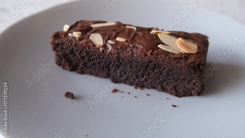 Smooth panning video showing a close-up of a rich, moist chocolate brownie slice topped with almond flakes, served on a white plate.
