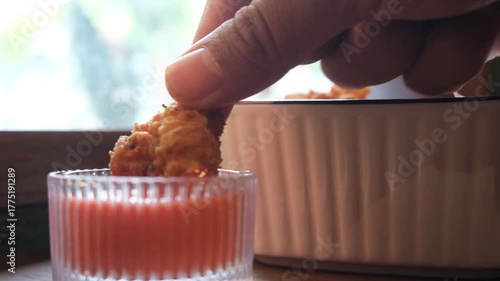 A hand dipping a crispy fried chicken piece into red dipping sauce, with a bowl of chicken in the background.