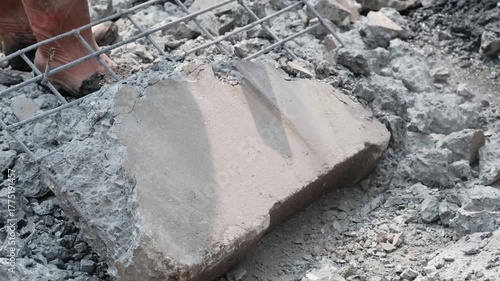 a construction worker using a sledgehammer to break a reinforced concrete slab with exposed rebar on a rubble-covered ground.  