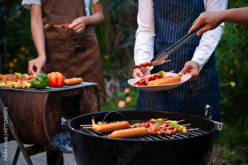 An Asian woman with friends grilling BBQ skewers, corn, and sausages outdoors. They toast with wine amid glowing string lights, creating a joyful and warm evening barbecue atmosphere