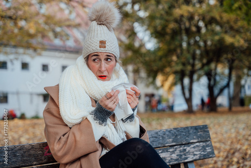 Woman sitting on park bench holding tissue sneezing in autumn air suffering from seasonal flu or allergy outdoors