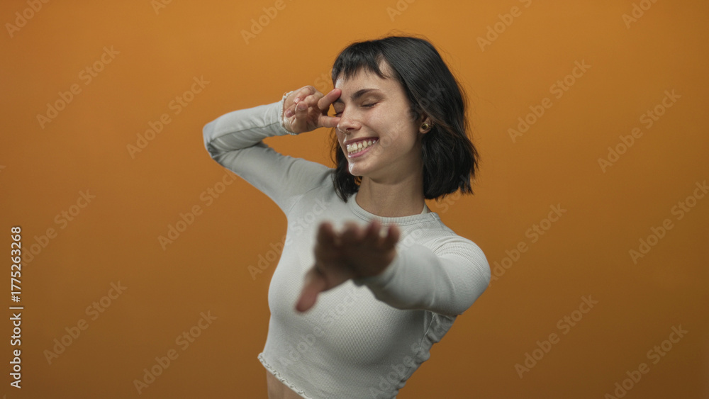Fototapeta premium Smiling young hispanic woman making peace sign near eye in studio against orange wall; serenity joy.