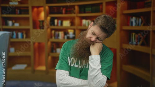 Man with long beard wearing volunteer shirt rests his head on closed fist and closes his eyes in library; fatigue.