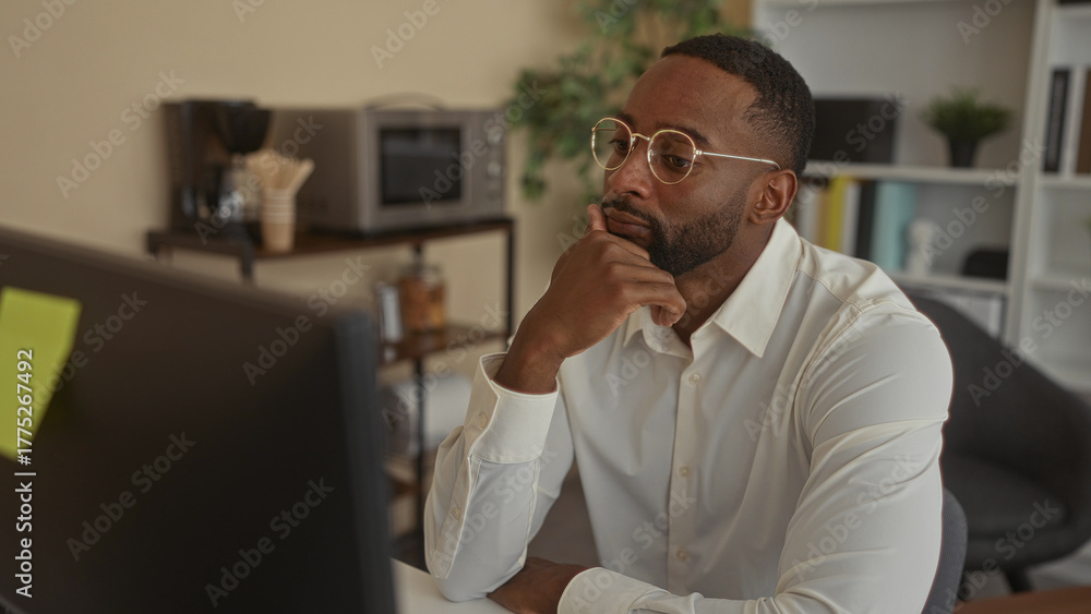 Fototapeta premium Man wearing white shirt and round glasses rests chin on hand at office desk while studying a monitor; pensive concentration.