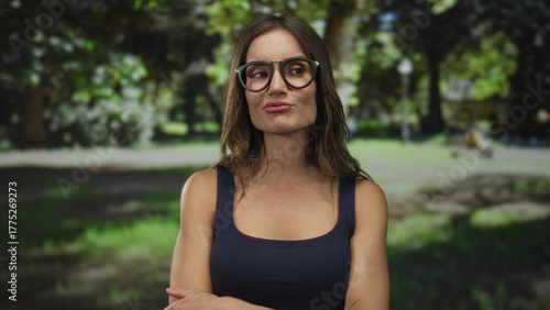 Woman wearing glasses and navy tank top purses lips and holds hand to head in green forest; confusion doubt uncertainty.