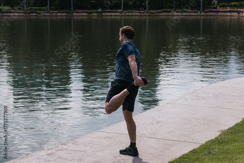 Man stretching quadriceps leg during an outdoor fitness workout by the lake, focusing on health and well-being
