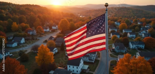 Fototapeta Naklejka Na Ścianę i Meble -  Aerial view of US flag waving proudly over small American town. Houses line quiet streets among vibrant autumn foliage. Warm golden sunset light illuminates rolling hills. Peaceful community embodies