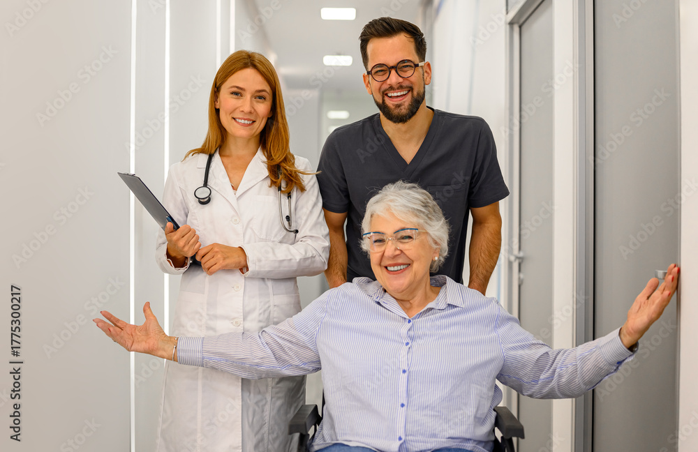 Obraz premium Portrait of confident doctors pulling cheerful senior patient sitting in wheelchair along hospital corridor