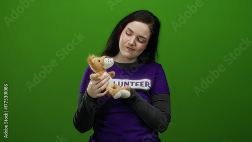 Woman volunteer with brunette hair holding stuffed toy against green background wearing purple shirt symbolizing compassion and community service.