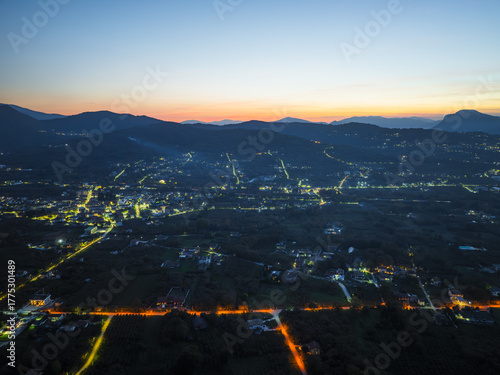 Aerial view of a tranquil town nestled amidst rolling hills, illuminated by the warm glow of streetlights against the deepening twilight, Santo Stefano del Sole, Irpinia, Campania, Italy.