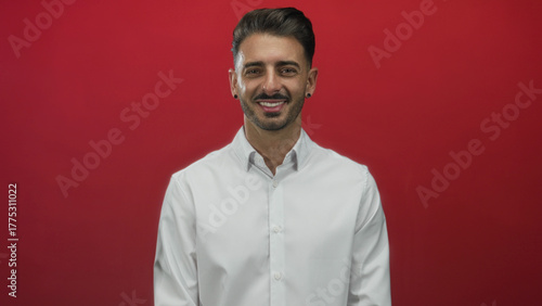 Quadro em tela Hispanic man face winking with toothy smile and confident gaze wearing white shirt in red studio; playful charm cheerful