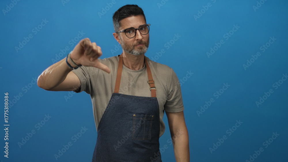 Fototapeta premium Man showing thumbs down gesture wearing denim apron in blue studio; disapproval critique feedback review.