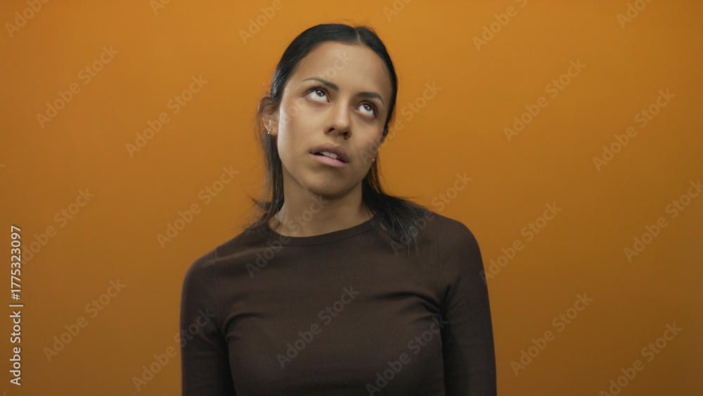 Fototapeta premium Woman expressing boredom over isolated orange wall embodying neutral emotion and casual style featuring young hispanic female in brown sweater with blank background wall setting.
