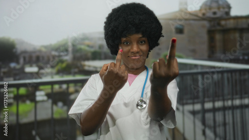Фотография Woman wearing white medical scrubs and blue stethoscope flips both middle fingers against an ancient roman building outdoors; defiance