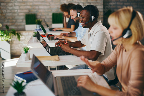Smiling African American male customer service agent works enthusiastically on his laptop and headset among diverse colleagues in a modern, busy call center office
