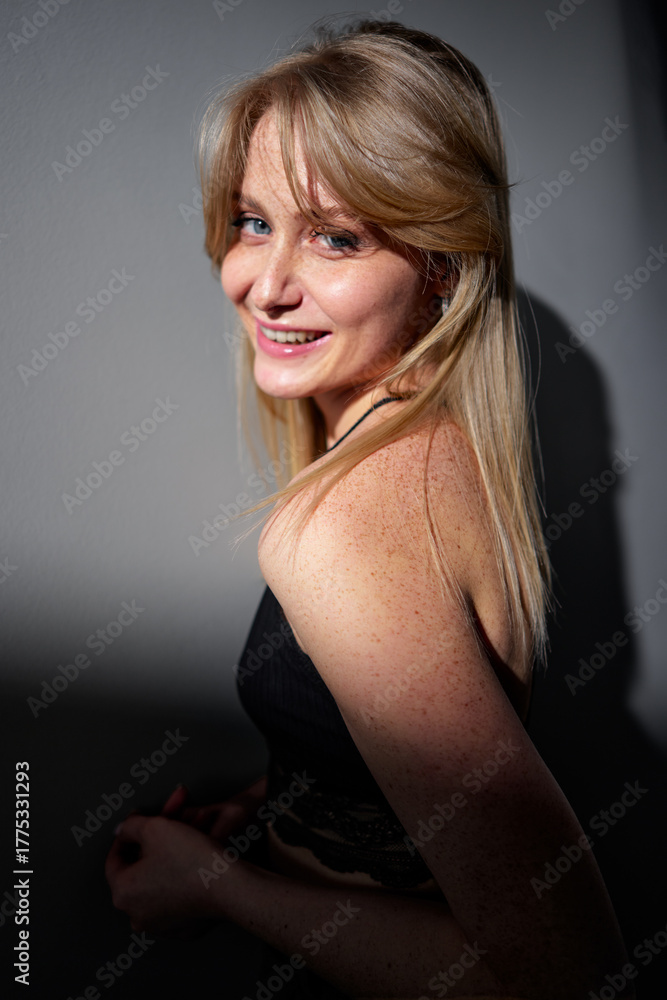 Naklejka premium A cheerful blonde woman with freckles smiles confidently, standing in natural light with shadow play creating an artistic effect. Her positive demeanor conveys a sense of joy and authenticity.
