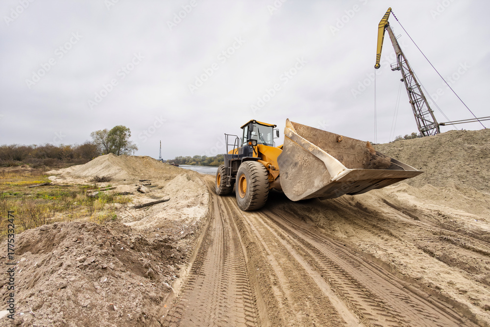 Fototapeta premium A giant loader works diligently, shifting soil along a riverbank, surrounded by mounds of dirt under a cloudy sky, signaling ongoing construction activity