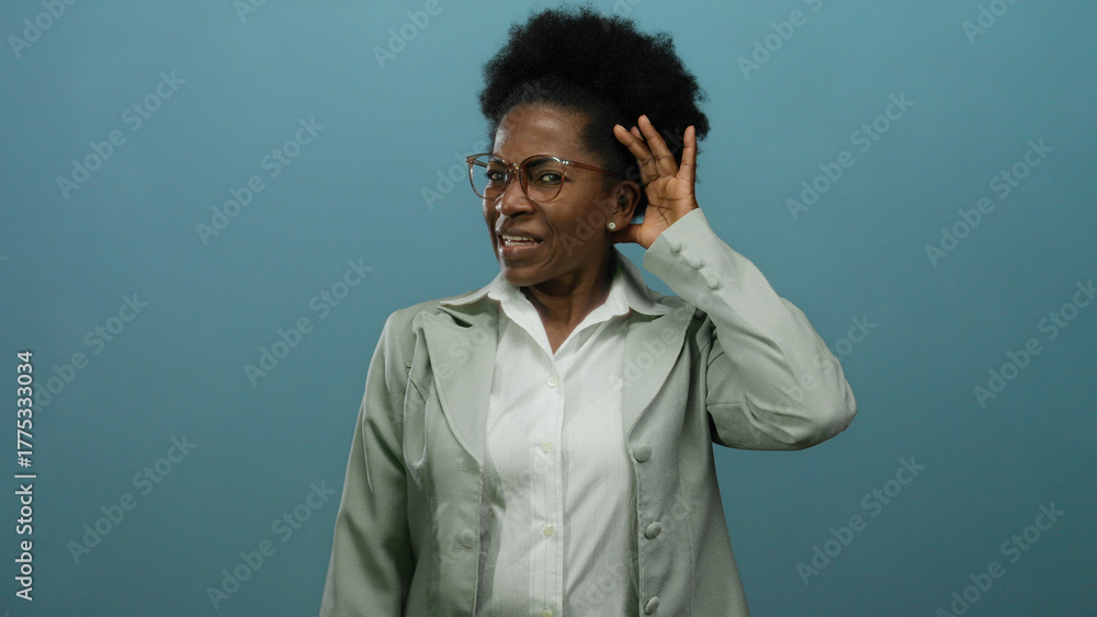 Fototapeta premium Woman listening intently with hand to ear against an isolated blue background, showcasing a thoughtful and curious expression in a business suit and glasses.
