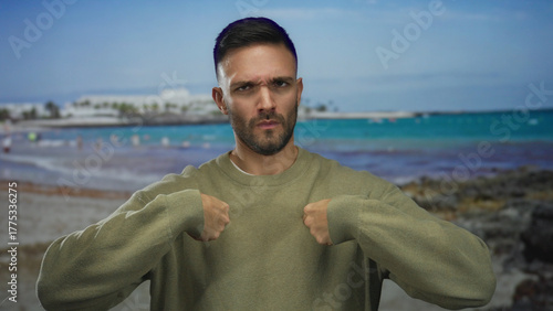 Hispanic man on beach with sweater gestures confidently by seashore, capturing a sunny outdoor scene with sea in background.