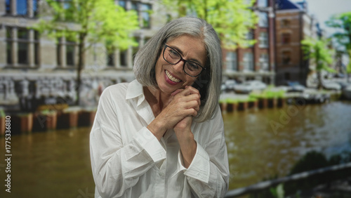 Canvastavla Woman smiling with hands clasped, glasses visible, standing by canal and historic building in amsterdam; serenity