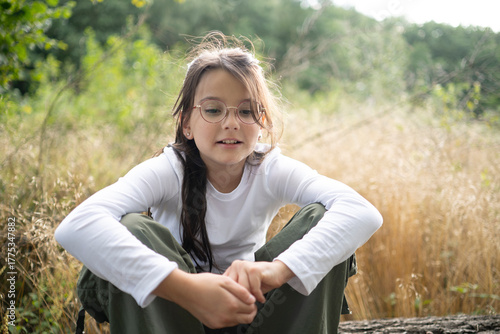 A smiling girl in glasses sitting in the forest, looking down thoughtfully with a gentle expression. Blurred golden grass and sky in the background, soft natural light, copy space. 