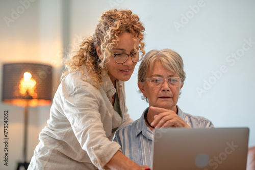 Smiling blonde curly woman helping an older woman use a laptop at home. The image represents intergenerational connection, technology learning, and family bonding
