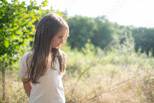 A cute little girl with long loose hair standing in the forest, turned half sideways and looking away. Blurred golden grass and trees in the background, warm natural light, copy space. 