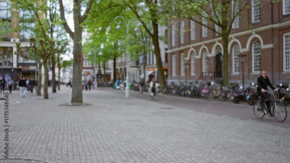 Obraz premium Blurred view of european city street with people walking and cycling on cobblestone path alongside historic buildings and green trees, creating a bokeh effect.
