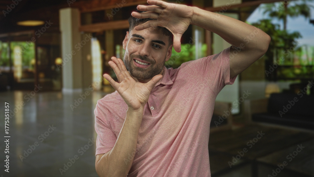 Obraz premium Smiling hispanic man in pink shirt framing hands to shape a view in hotel building; creative vision.