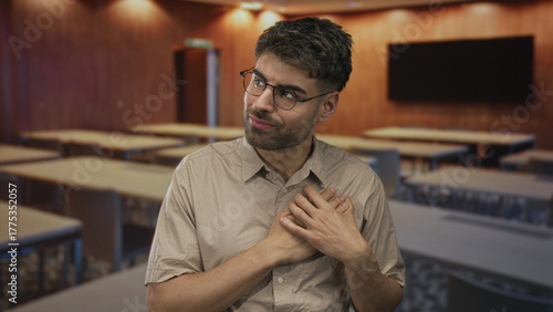 Foto Man clutching chest at wooden desk amid rows of desks in bright classroom indoors; anxiety worry concern tension