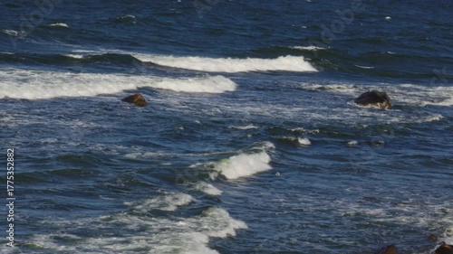 Ocean Waves Crashing Over Rocks on a Sunny Day, Creating White Foam. Beautiful Seascape with Deep Blue Water and Rocky Coastline in Natural Sunlight with Rolling Surf