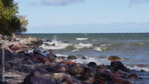 Waves crashing on the rocky Baltic Sea shoreline with autumn trees and colourful boulders. Coastal landscape showing a natural seascape with moss-covered stones and white foamy water splashing.