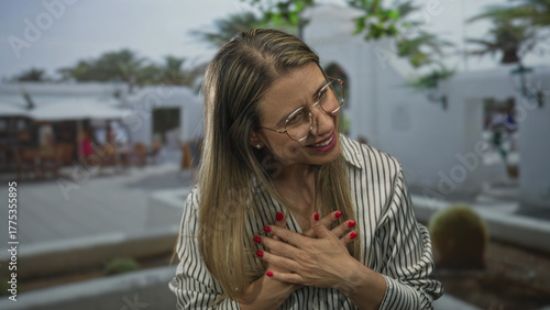 Tableau sur toile Middleaged blonde woman with glasses holding hands over chest on urban street; sincerity gratitude warmth