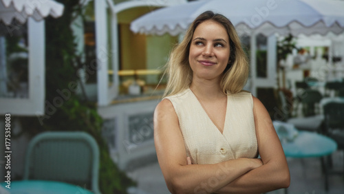 Woman with arms crossed smiling on restaurant terrace among outdoor tables and umbrellas; casual confidence. Free Download Woman with arms crossed smiling on restaurant terrace among outdoor tables and umbrellas; casual confidence. - GFXLoad