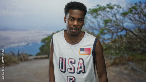 Φωτογραφία Young african american man wearing white usa basketball jersey places hand on chest in forest; pain