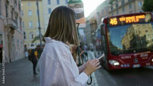 Photography Woman brunette tapping smartphone with fingers at a street bus stop beside rome buildings and a red bus; calm commute