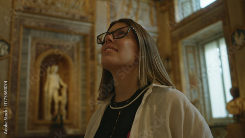 Bilde på lerret Young woman looking up face in ornate museum building, wearing glasses and necklace, admiring marble statue; awe curiosity
