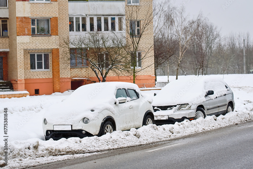 Fototapeta premium Cars parked on snowy roadside blocked by slush piles, impact of snowplow work during winter storm. Roadside parking zone in residential area with snow covered cars, aftermath of municipal snow removal